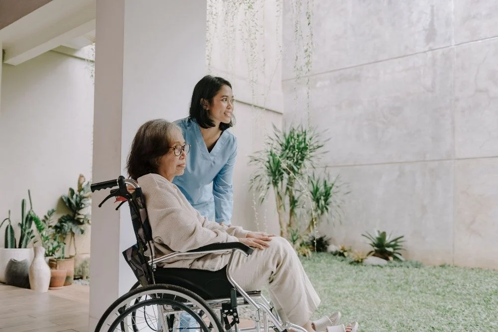 A caregiver pushes an elderly woman in a wheelchair. Webster & Garino on Indiana nursing home asset protection.