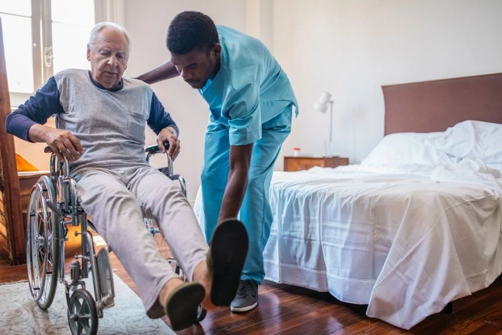 Nurse assists elderly man into wheelchair beside bed; Indiana nursing home cost planning context.