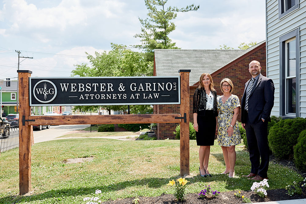 webster-legal-bottom-blog-image Three attorneys stand beside the Webster & Garino, LLC Attorneys at Law office sign outdoors on a sunny day.