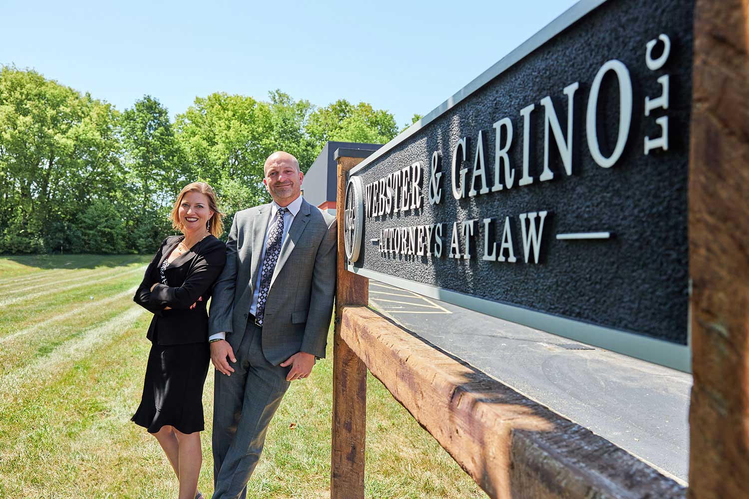 New-Team-Webster-and-Garino-No-Danika Two attorneys stand smiling beside a sign for a trusted Indiana law firm, with trees in the background.| Webster & Garino, LLC | Indiana Law Firm serving Hamilton County