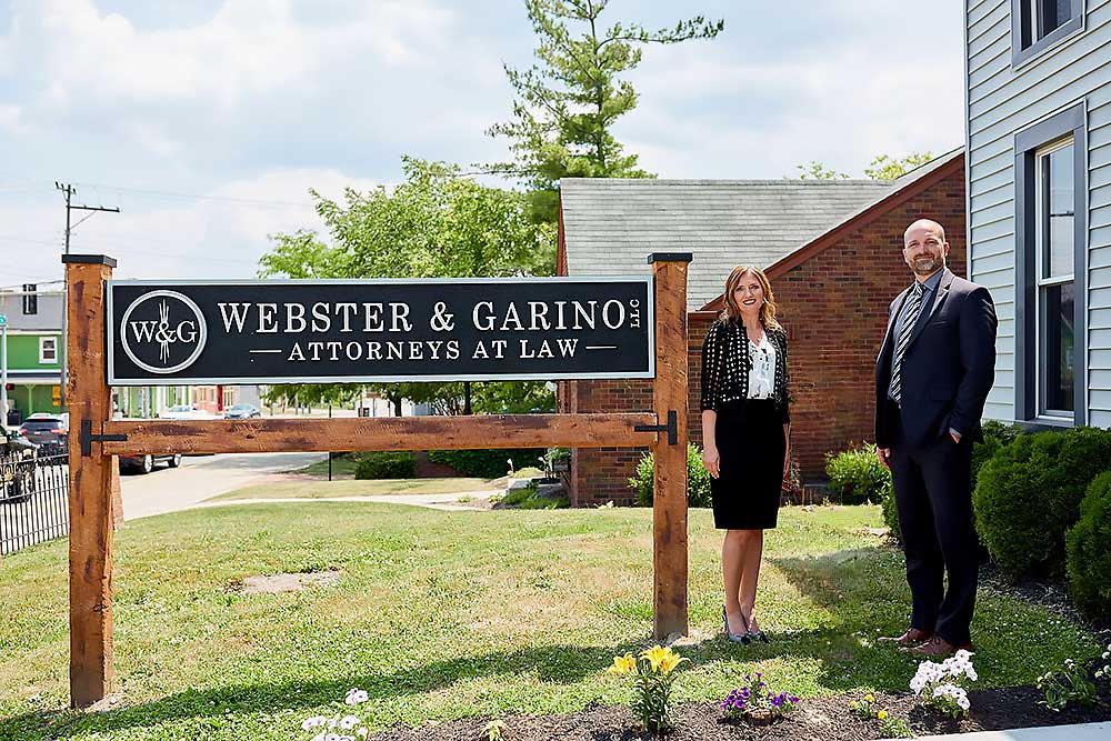Three attorneys stand beside the Webster & Garino, LLC Attorneys at Law office sign outdoors on a sunny day.